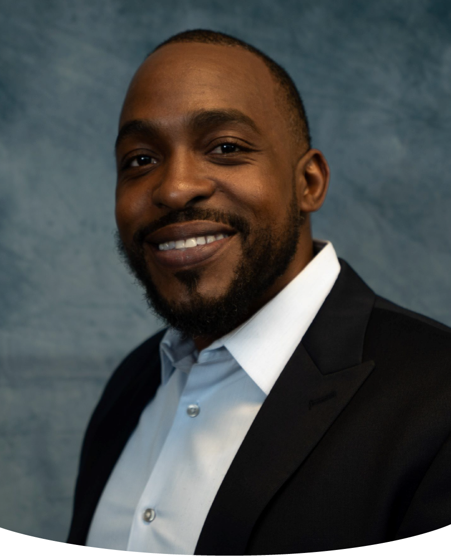 Smiling Black man in a suit jacket and button-down shirt, posing against a blue background.