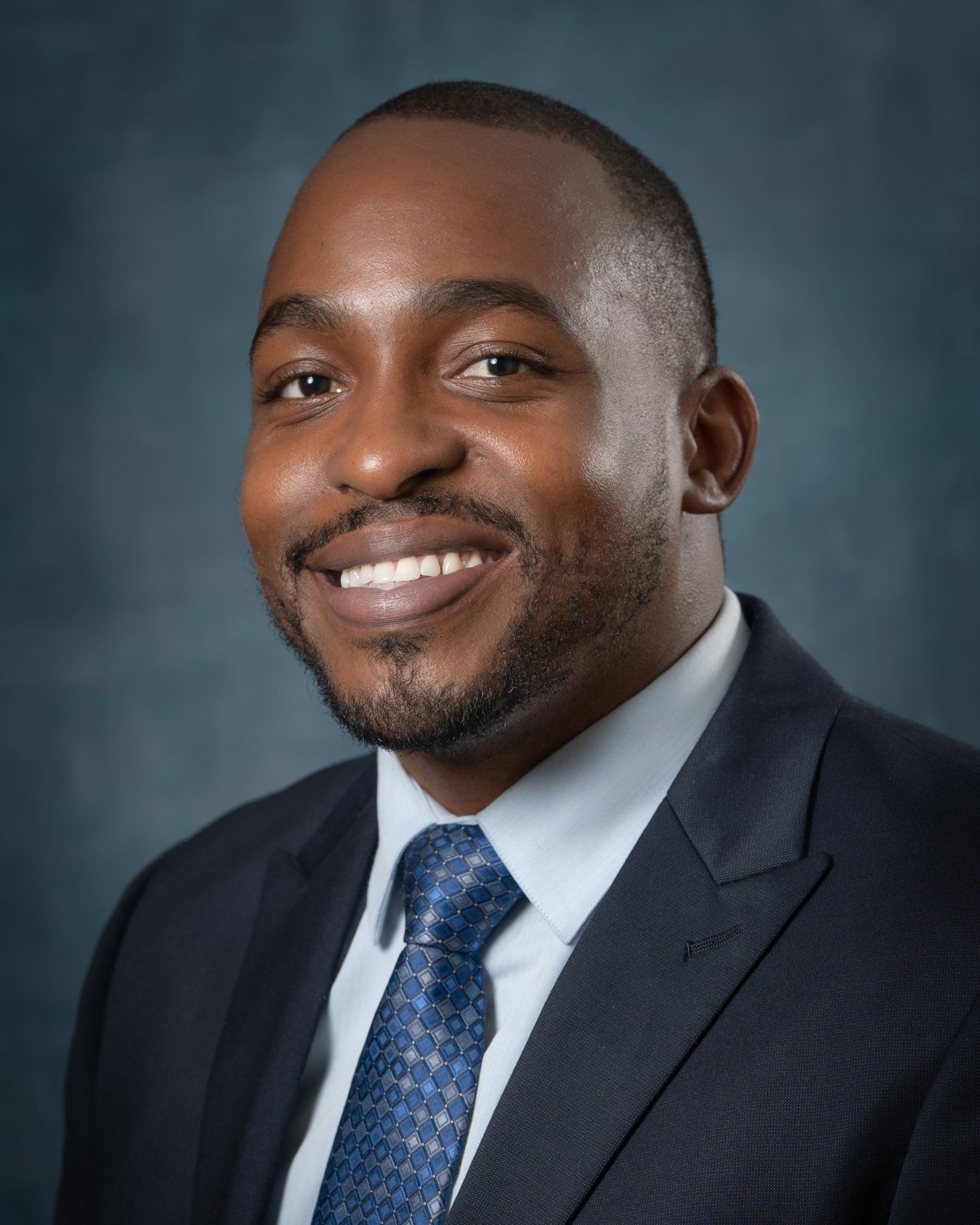 Smiling Black man in suit and tie, against a blurred dark blue background.