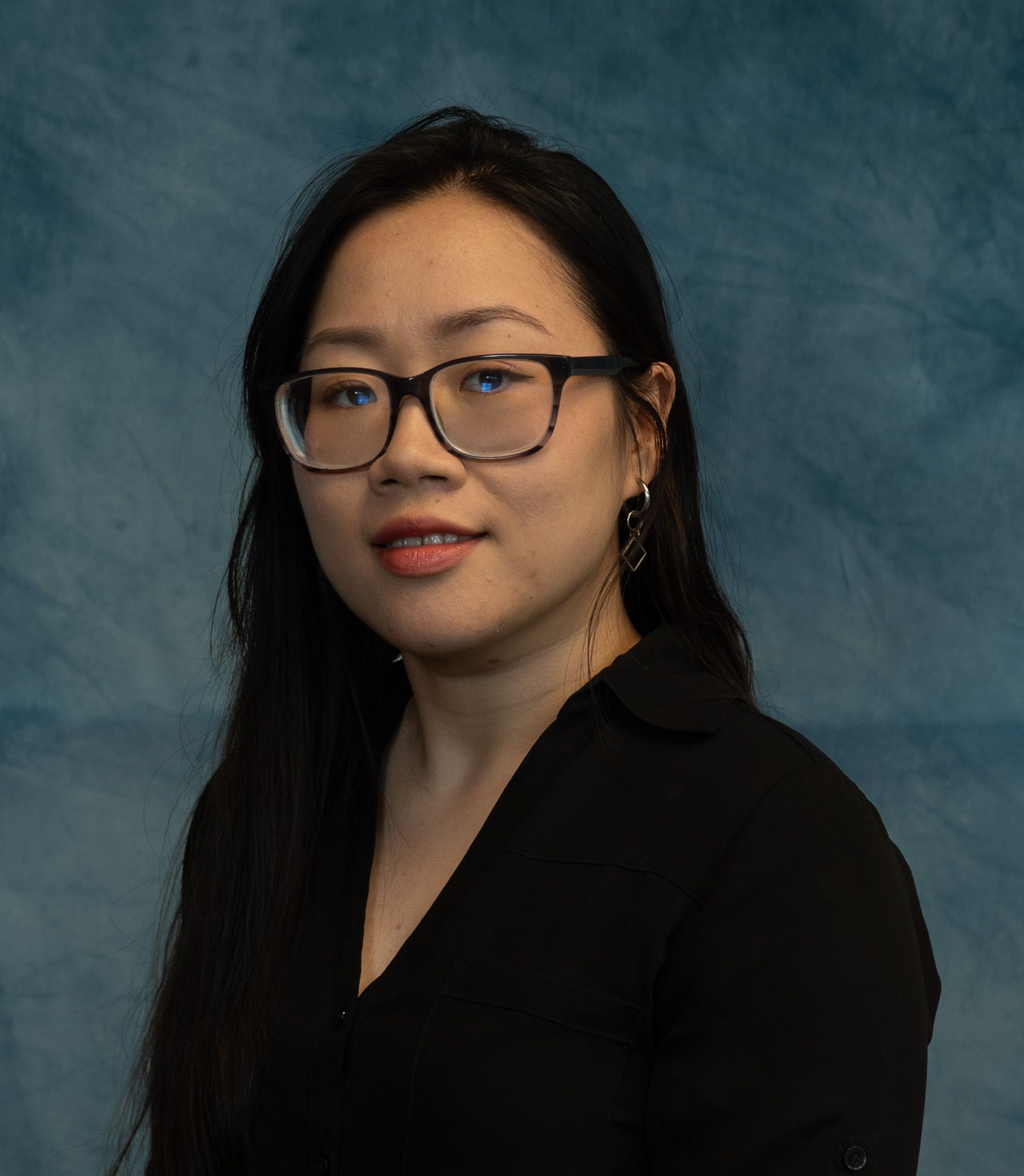 Woman with glasses, dark hair, black shirt, smiling; blue backdrop.