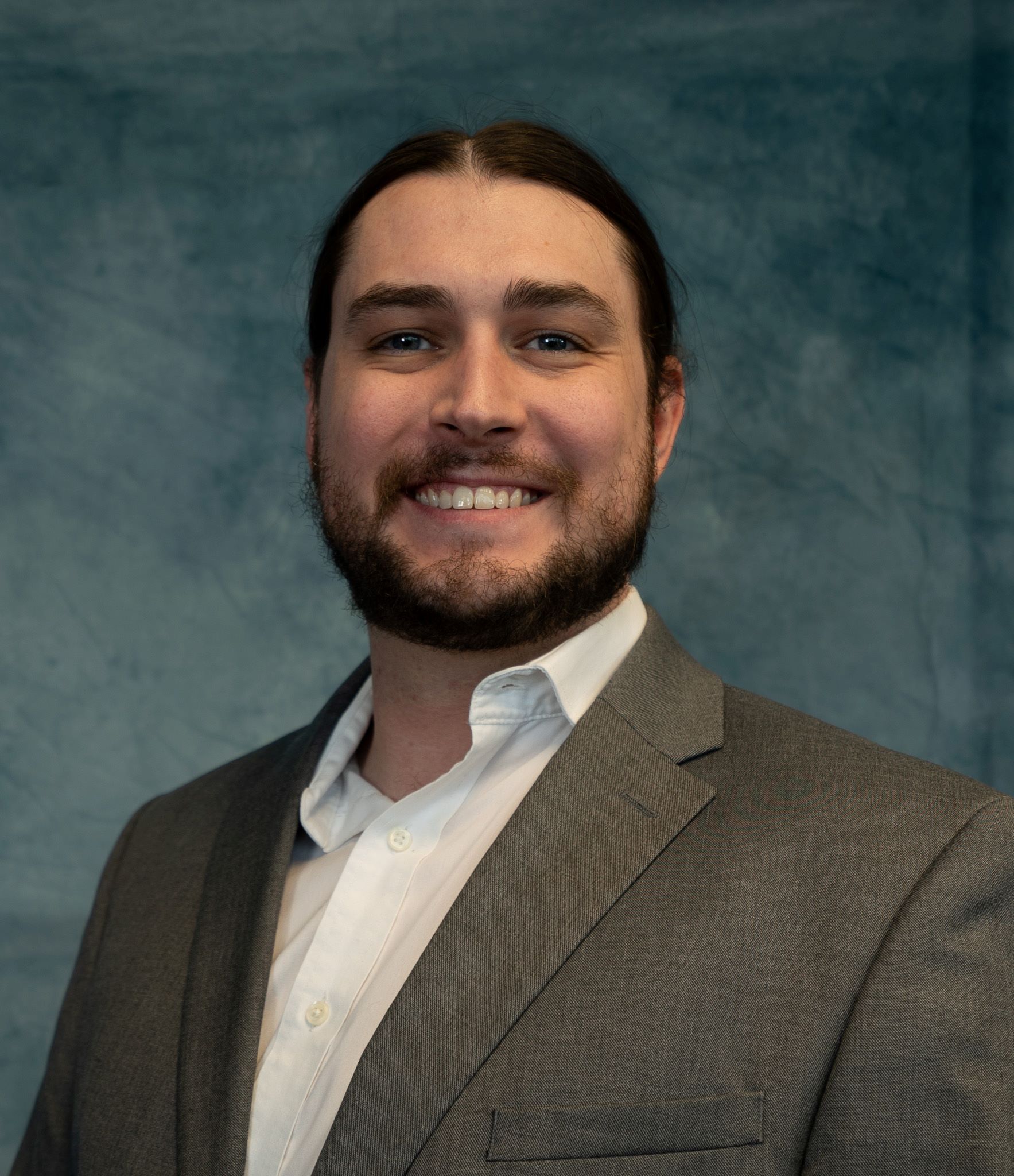 Man with dark hair and beard smiles wearing a gray suit jacket and white shirt.