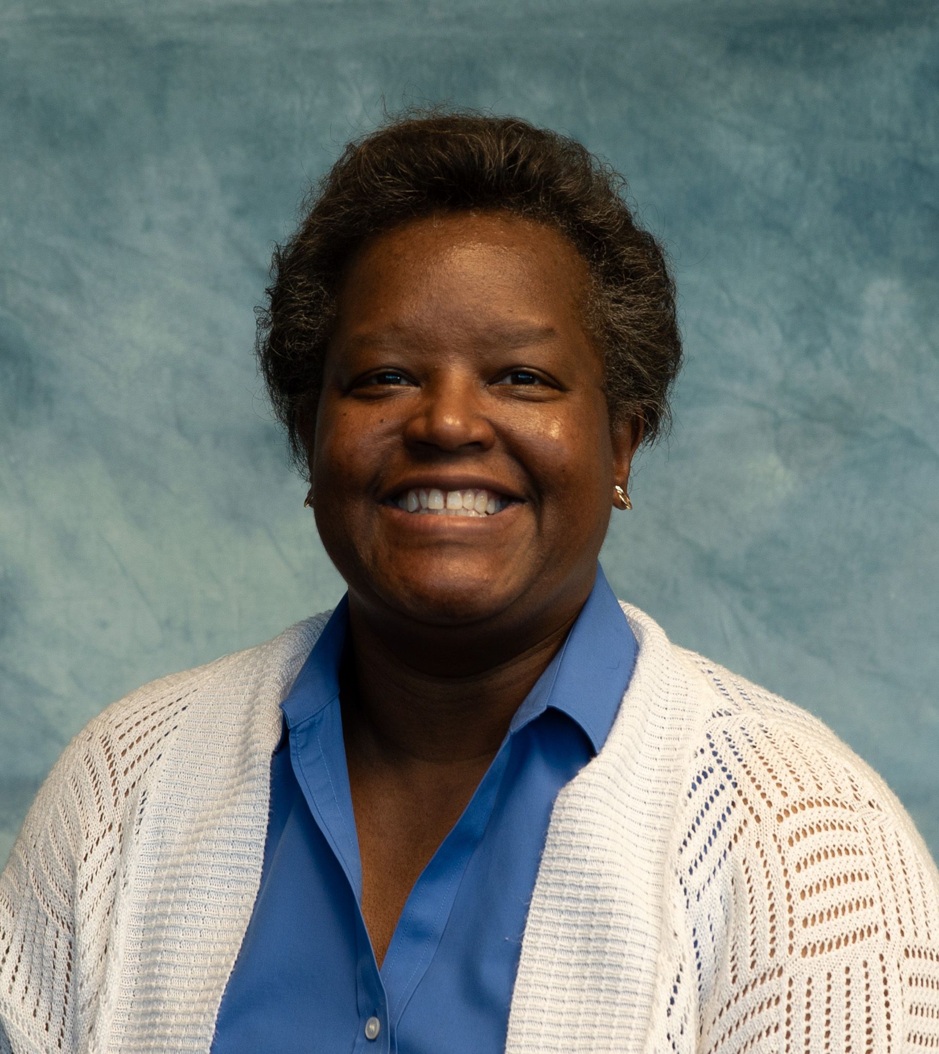 Smiling Black woman wearing a blue shirt and white cardigan in front of a blue backdrop.