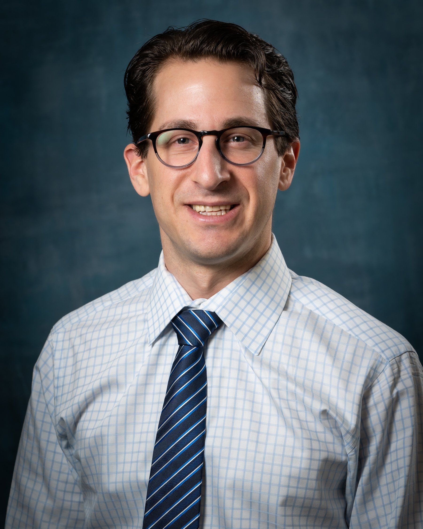 Man with glasses smiling, wearing a blue and white checkered shirt and tie. Dark blue background.