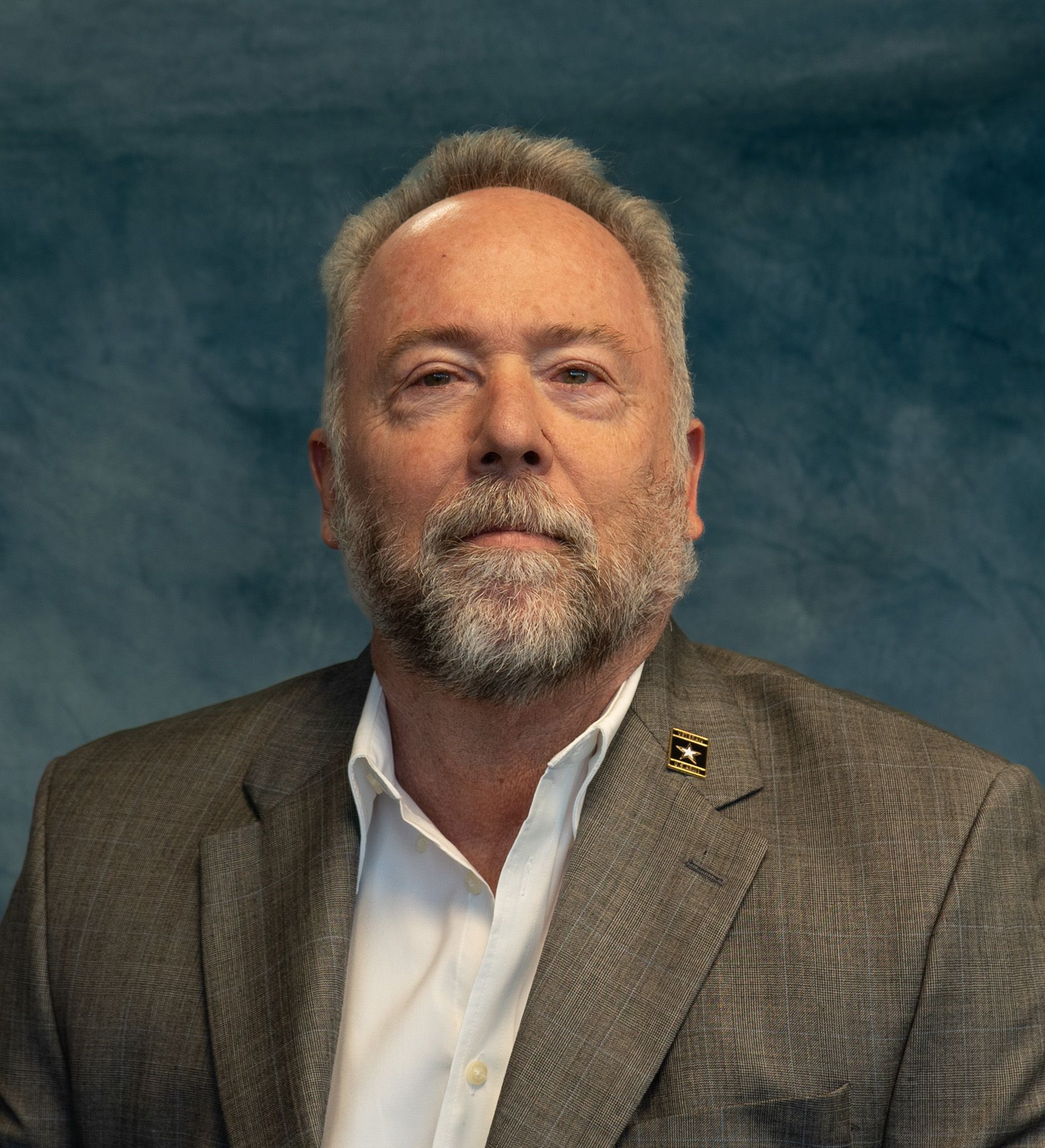 Man with gray hair and beard in a gray jacket and white shirt, looking forward. Blue background.