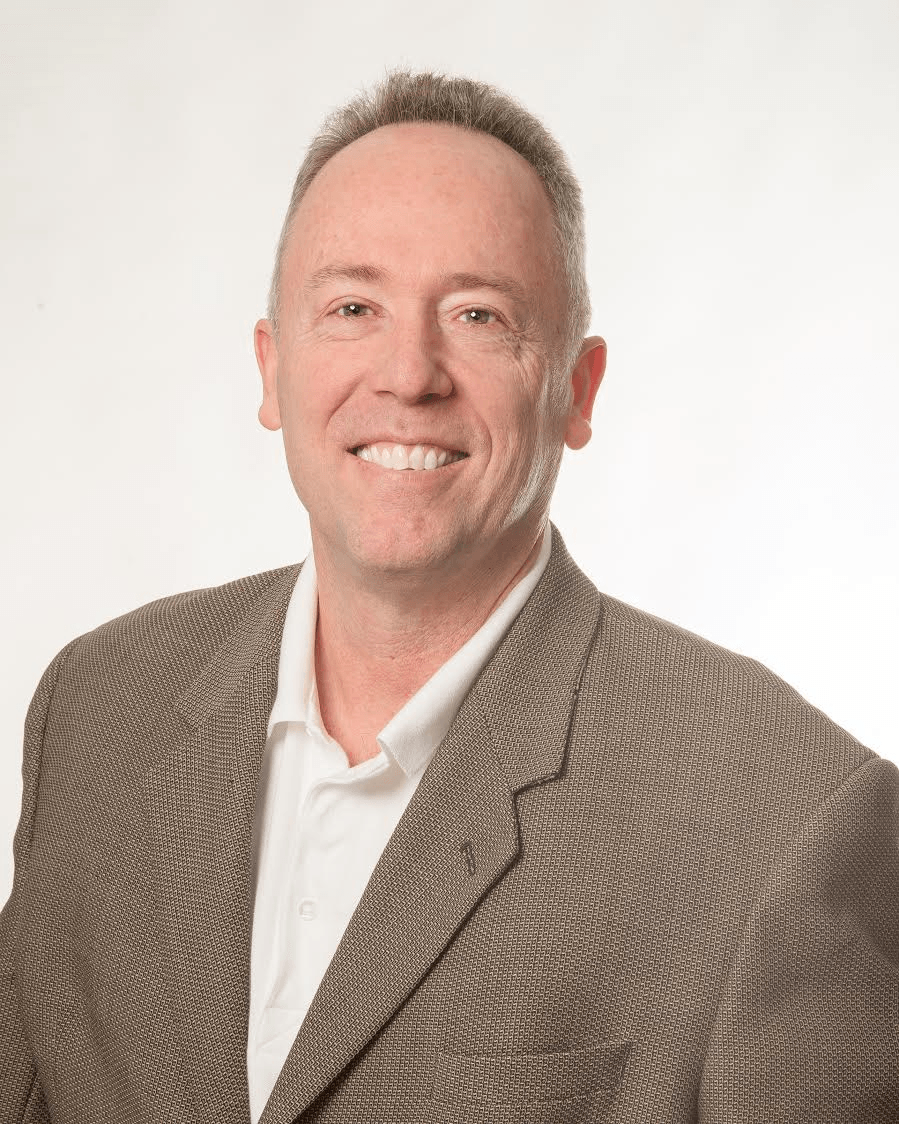 Man in a tan blazer smiles at the camera. White shirt, neutral background.