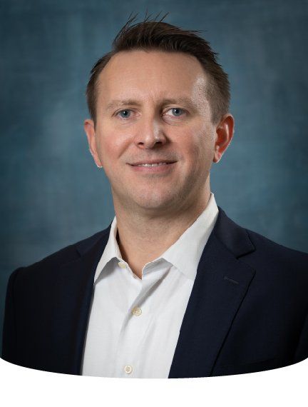 Man with short brown hair, blue eyes, smiling, wearing a white shirt and navy blazer, against a blue backdrop.