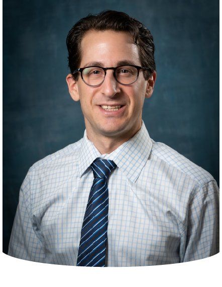 Man with glasses wearing a tie and button-down shirt, smiling, against a blue background.