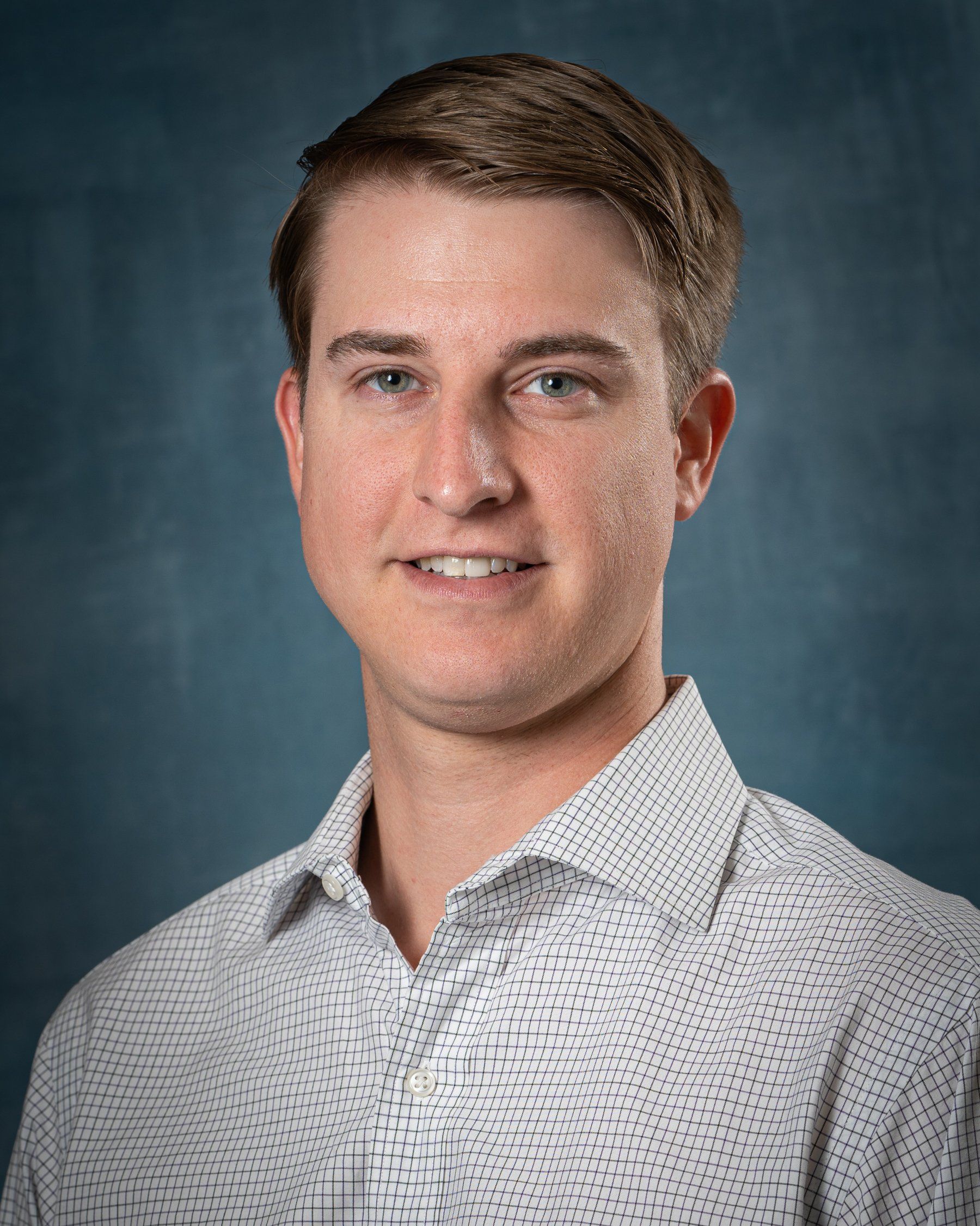 Smiling young man with light skin wearing a patterned button-down shirt, in front of a blue backdrop.