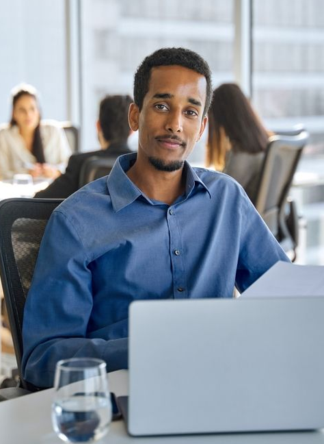 Man sitting at a desk with a laptop in a bright office, with coworkers blurred in the background