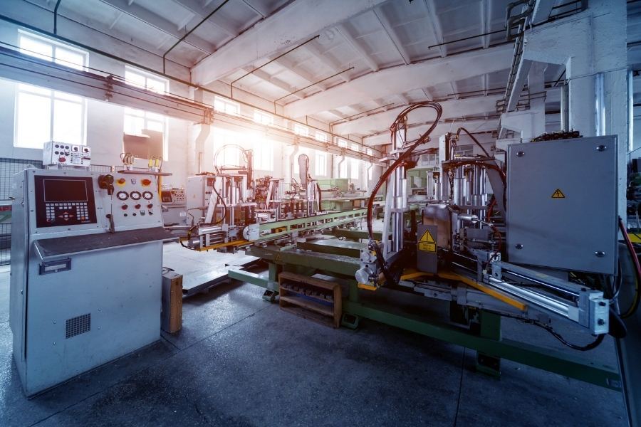 Factory interior with machinery; a control panel on the left. Bright lighting.