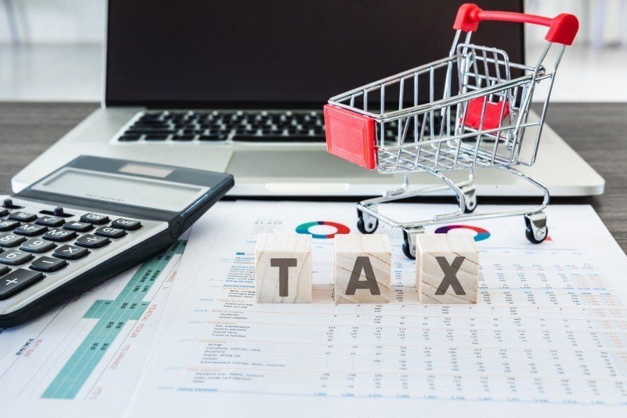 A laptop, calculator, and shopping cart sit on documents labeled with the word “TAX” on blocks.