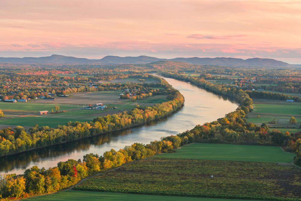 Wide aerial view of a river winding through autumnal trees and fields; distant mountains and pink sky.