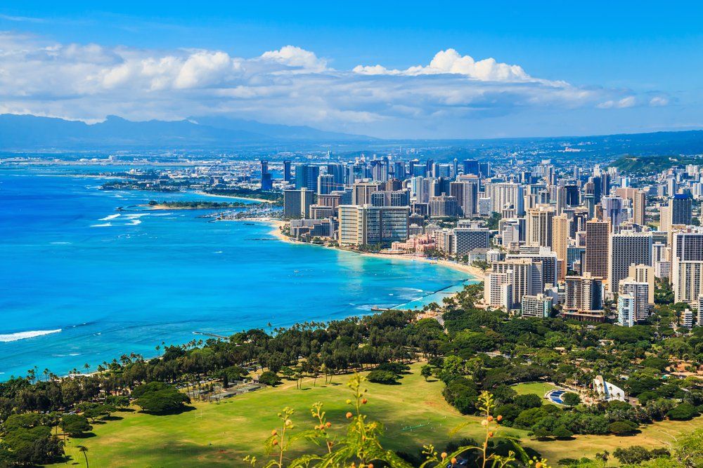 Coastal city view with turquoise water, buildings, and green landscape under a blue sky.