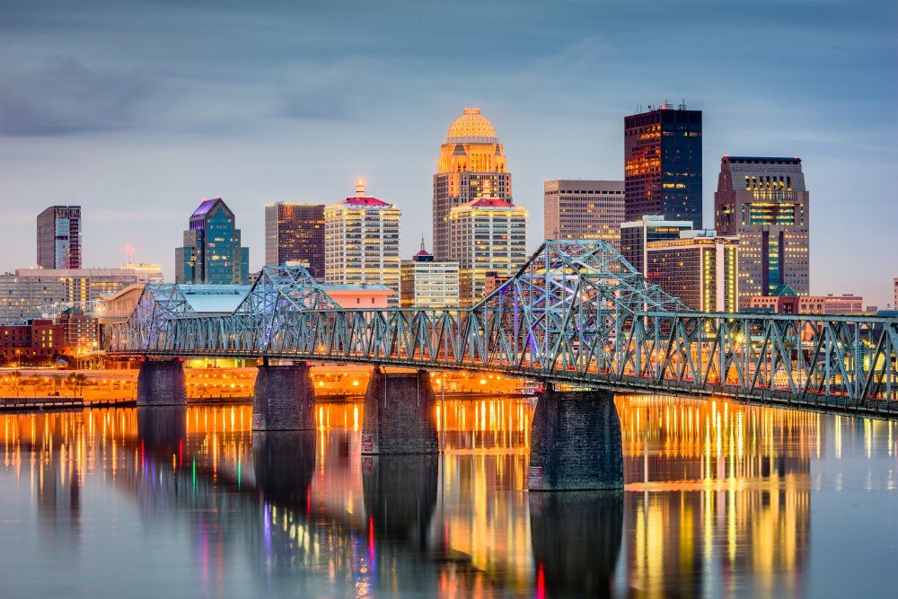 City skyline at dusk, featuring a bridge over calm water, with buildings illuminated by lights.