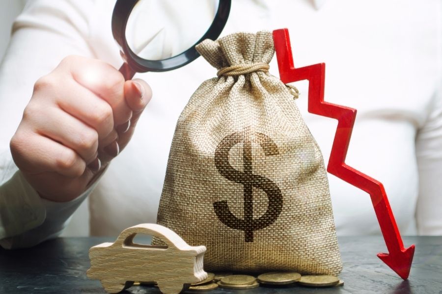 Person examining money bag with dollar sign, a downward red arrow, and a toy car.