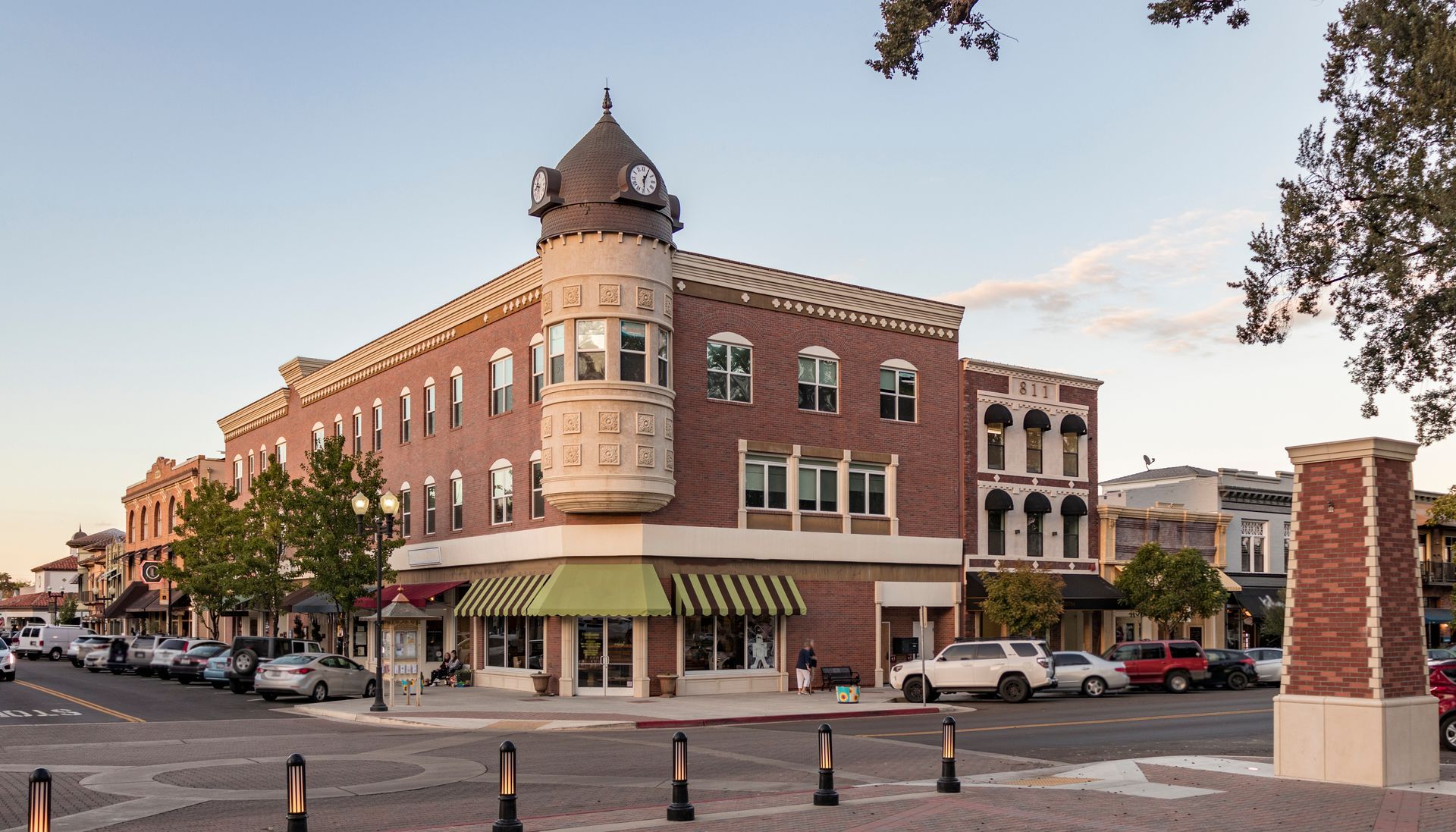 Brick building with a turret, storefront with striped awning, parked cars line the street.