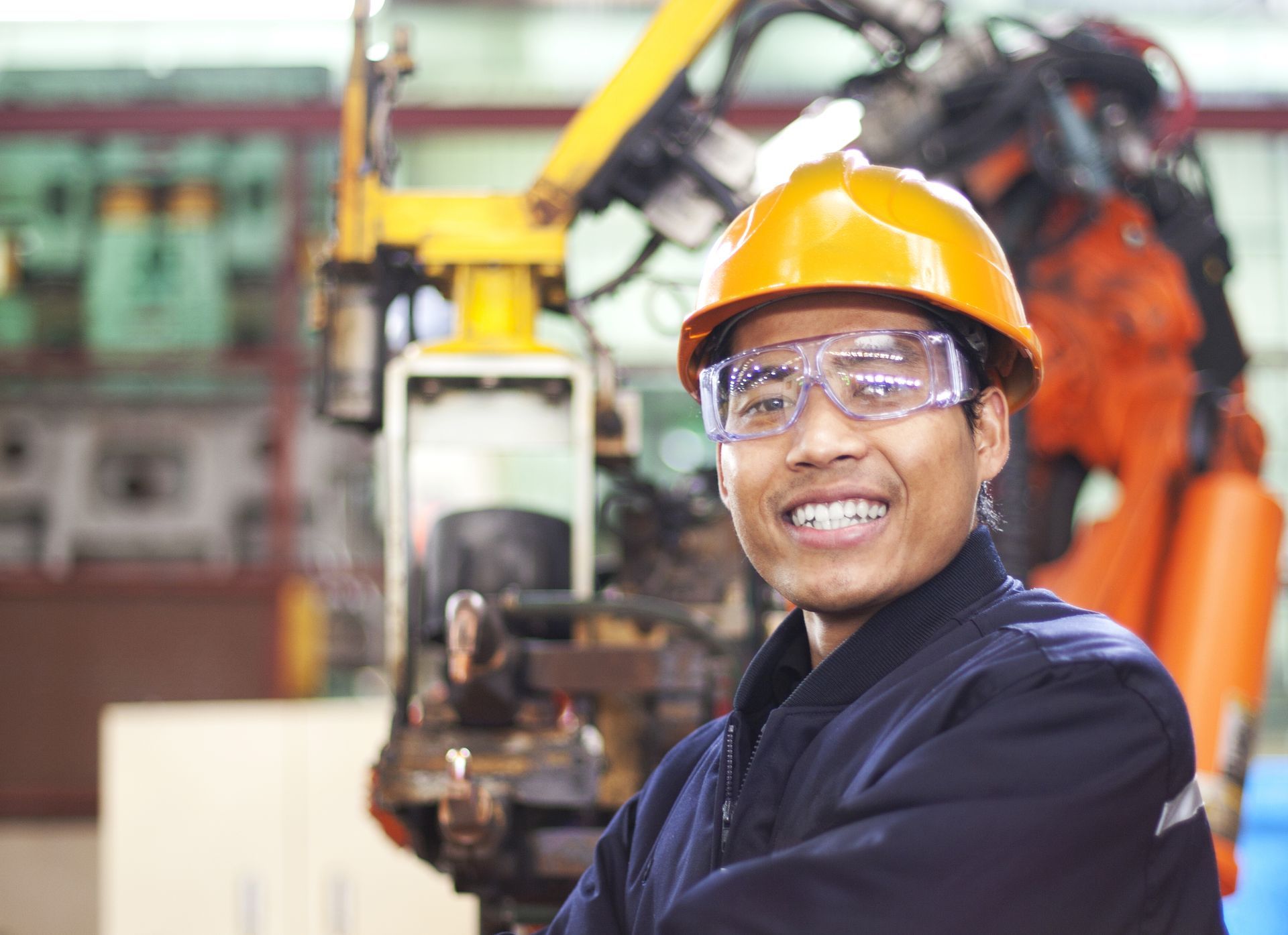 Smiling man in a yellow hard hat and safety glasses in a factory, near a robotic arm.