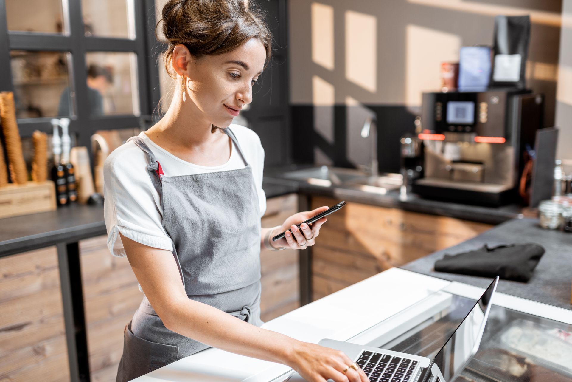 Woman in apron using a laptop and phone behind a cafe counter near a coffee machine.