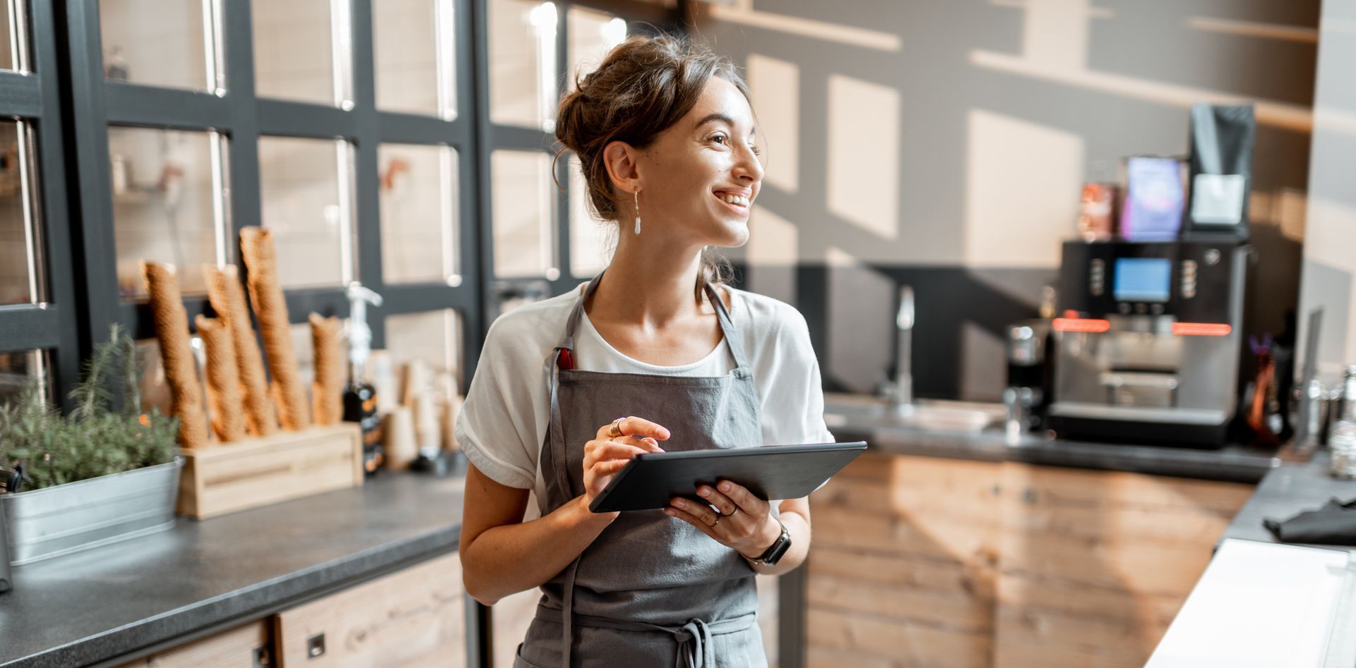 Woman in apron smiles while holding tablet in a cafe, near a counter with coffee machine.