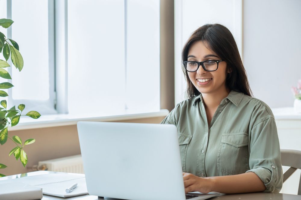 Woman with glasses smiles while using a laptop at a desk near a window with a plant.