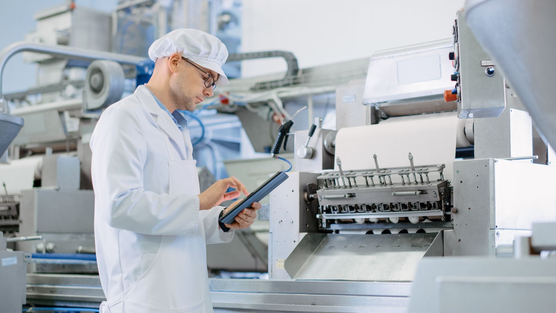 Man in lab coat inspects machinery with tablet in food processing plant.