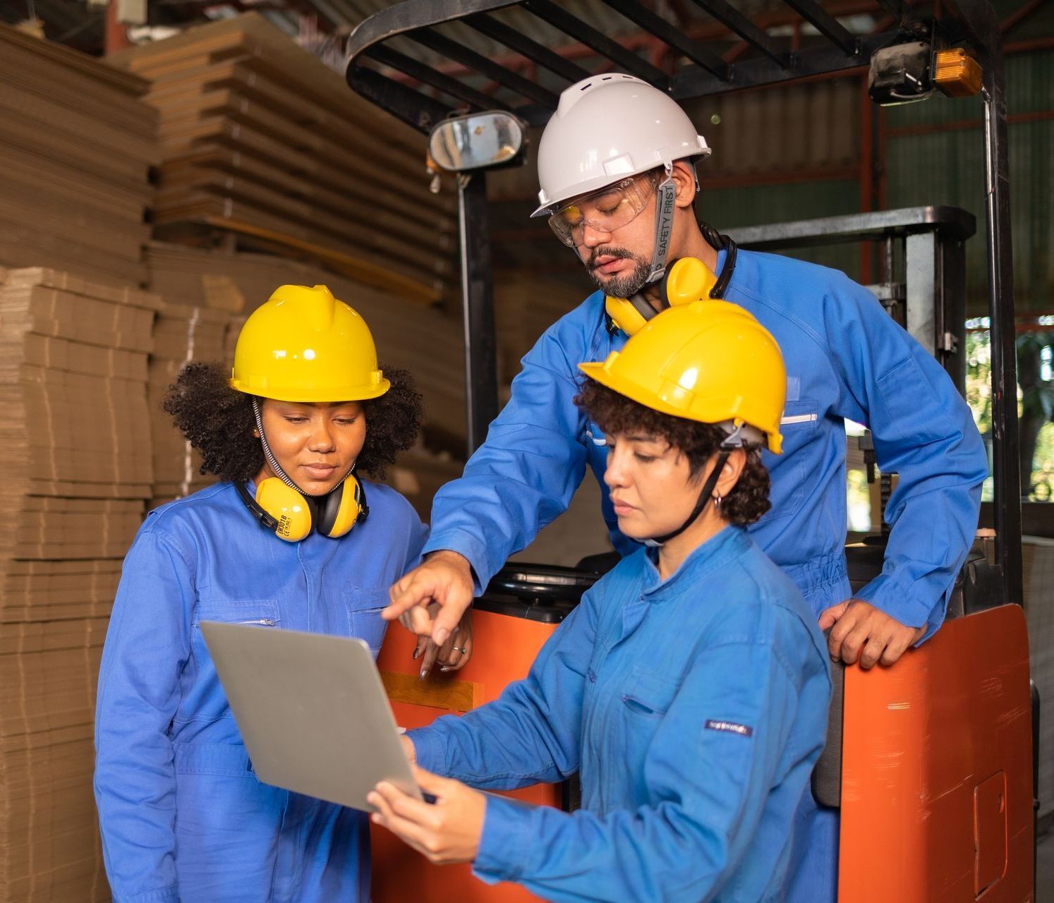 Three workers in a warehouse discussing a laptop. They wear hard hats and work uniforms near a forklift.