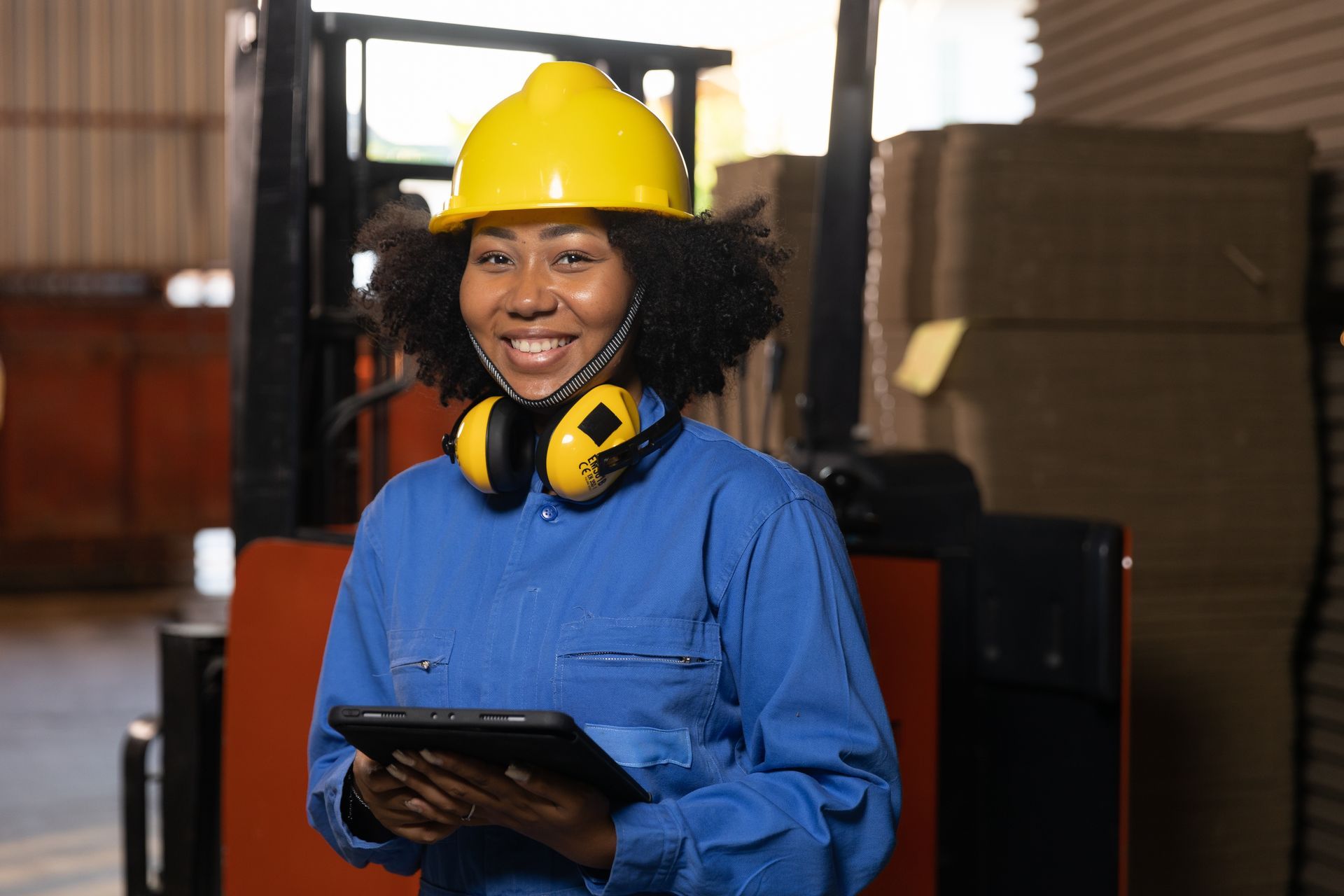 Woman in work clothes and yellow hard hat, holding tablet, smiles in warehouse near forklift.