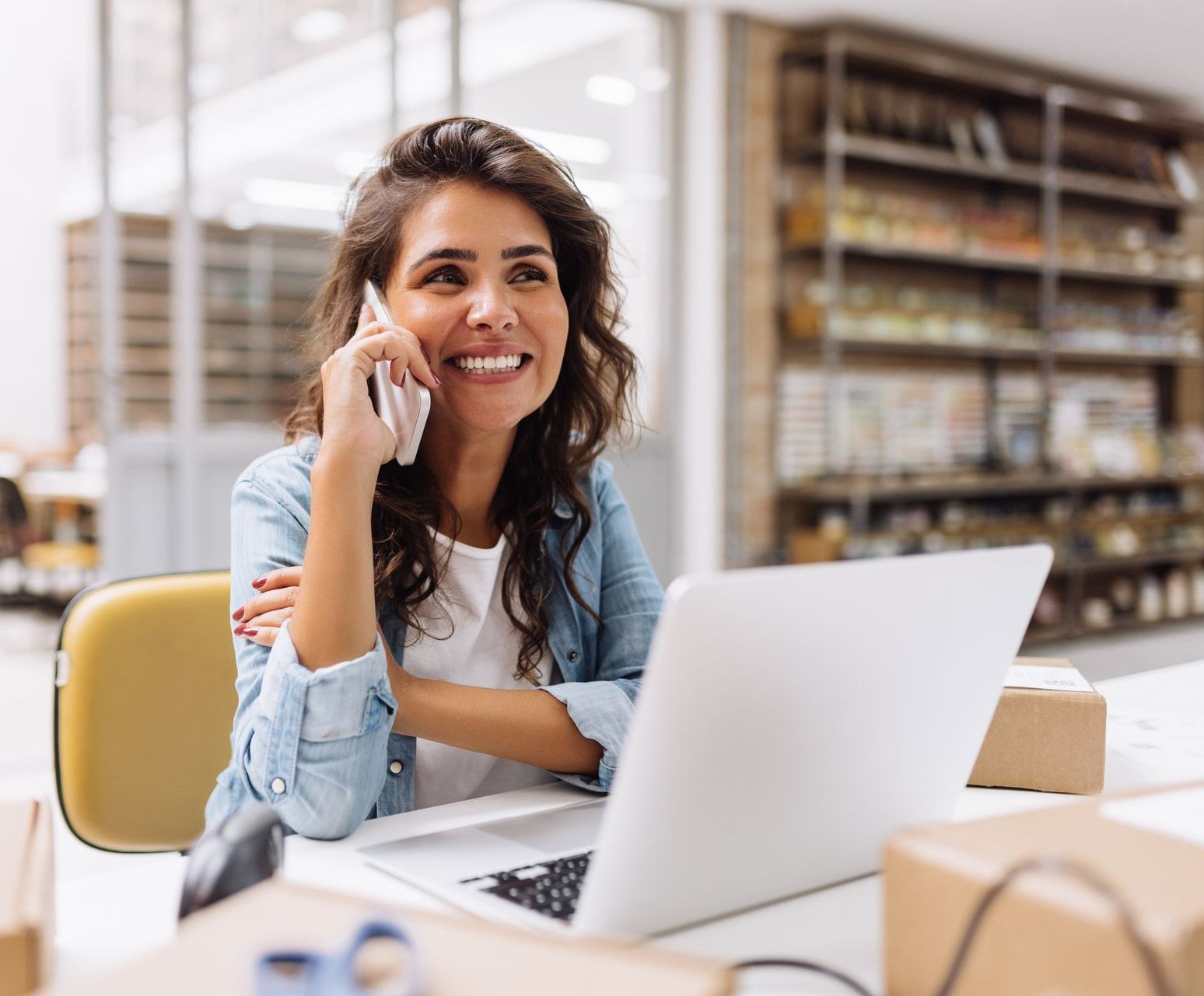 Woman smiling, talking on phone, working on laptop at desk; retail storage shelves in background.