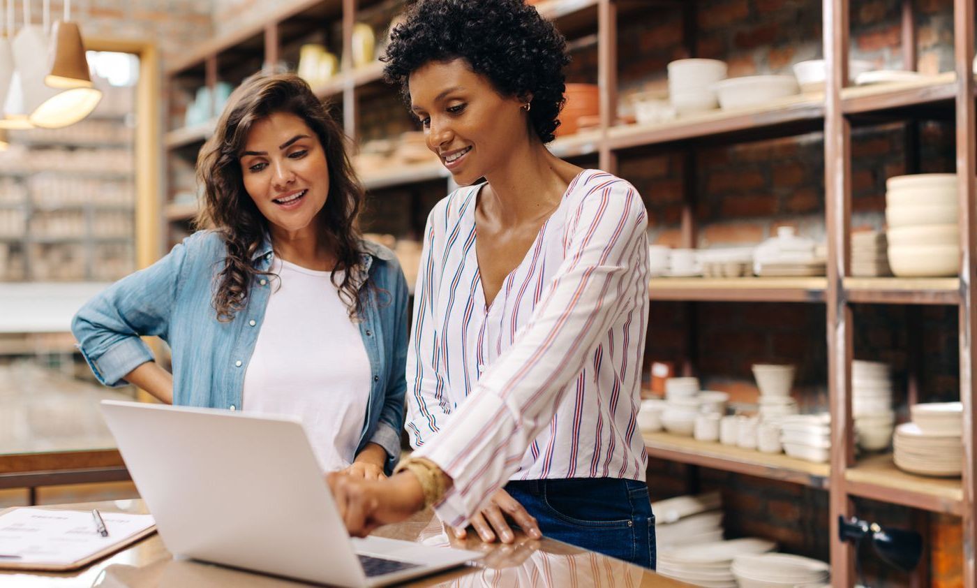 Two women look at a laptop in a pottery shop; one points, shelves of pottery in the background.