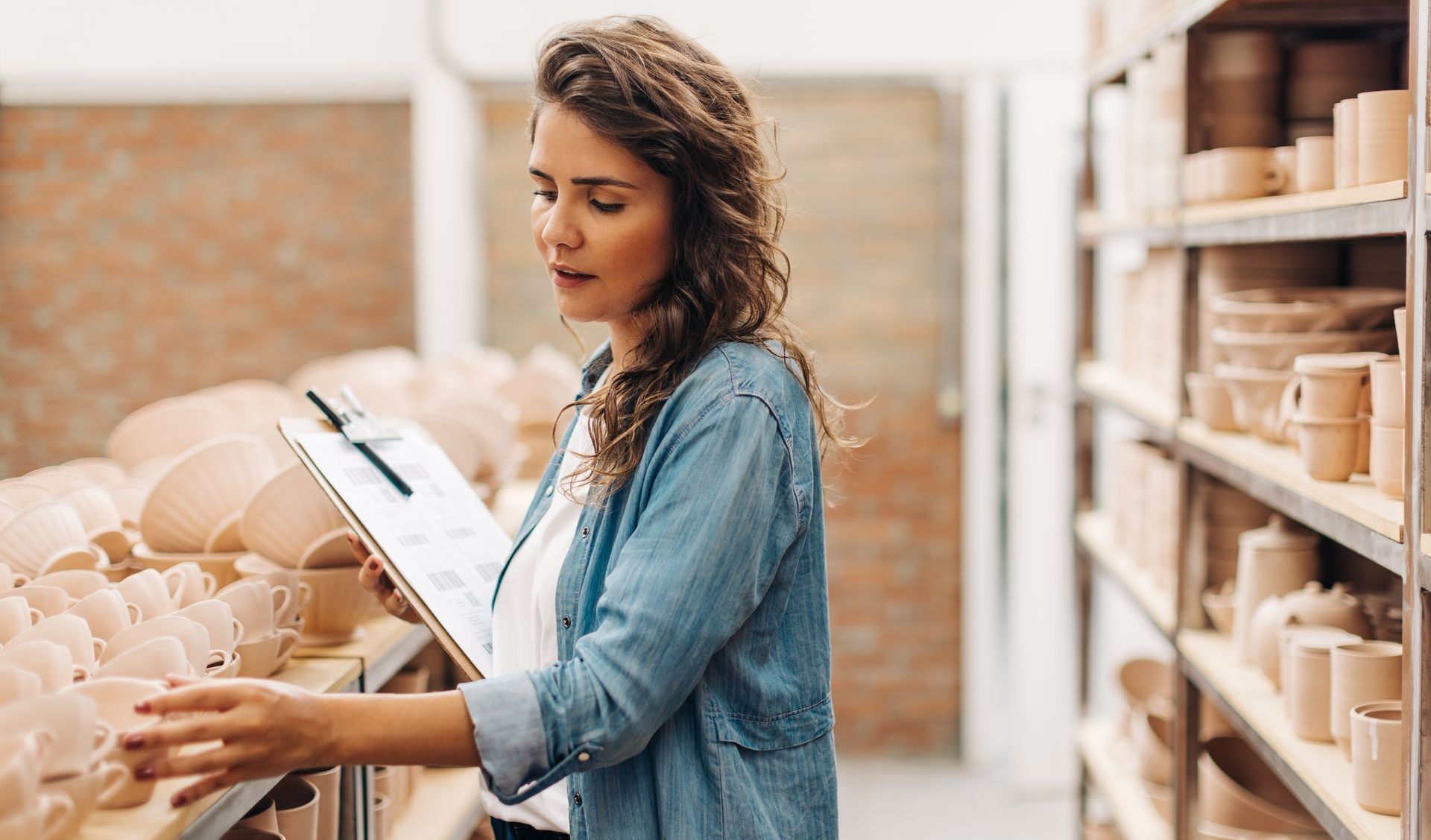 Woman in denim shirt inspecting pottery on shelves, holding clipboard.