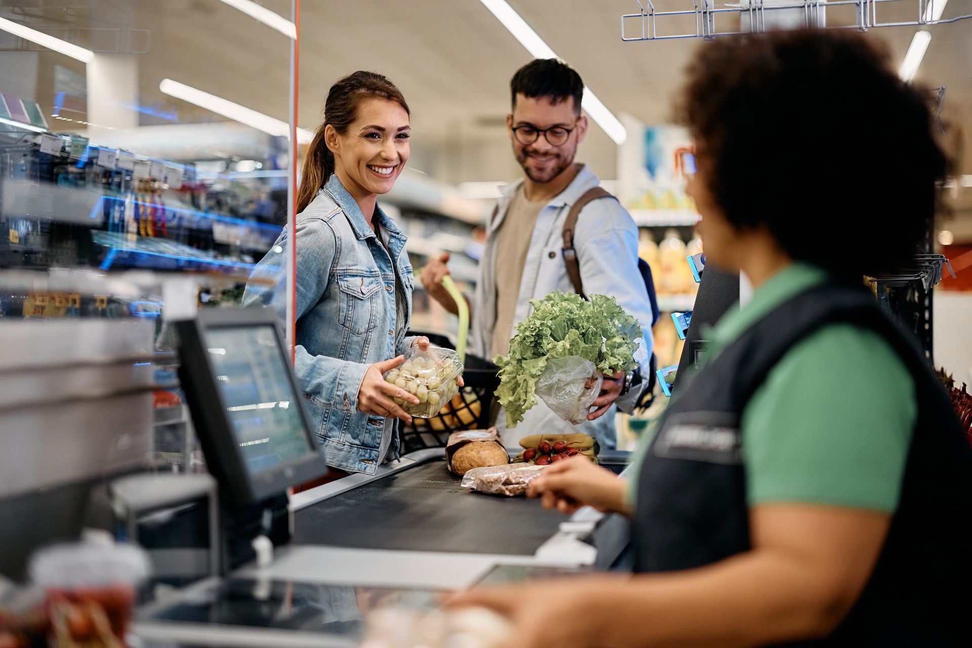 A couple at a supermarket checkout, smiling while handing items to a cashier.