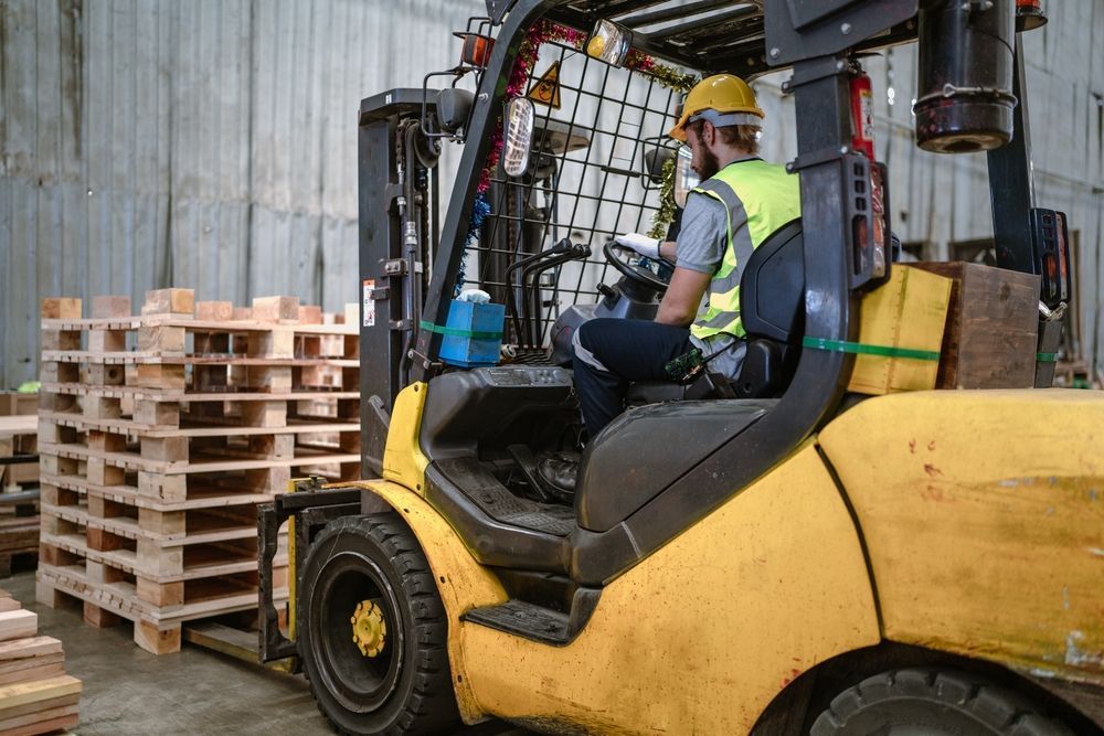 Forklift operator in a warehouse, wearing safety gear, near wooden pallets.