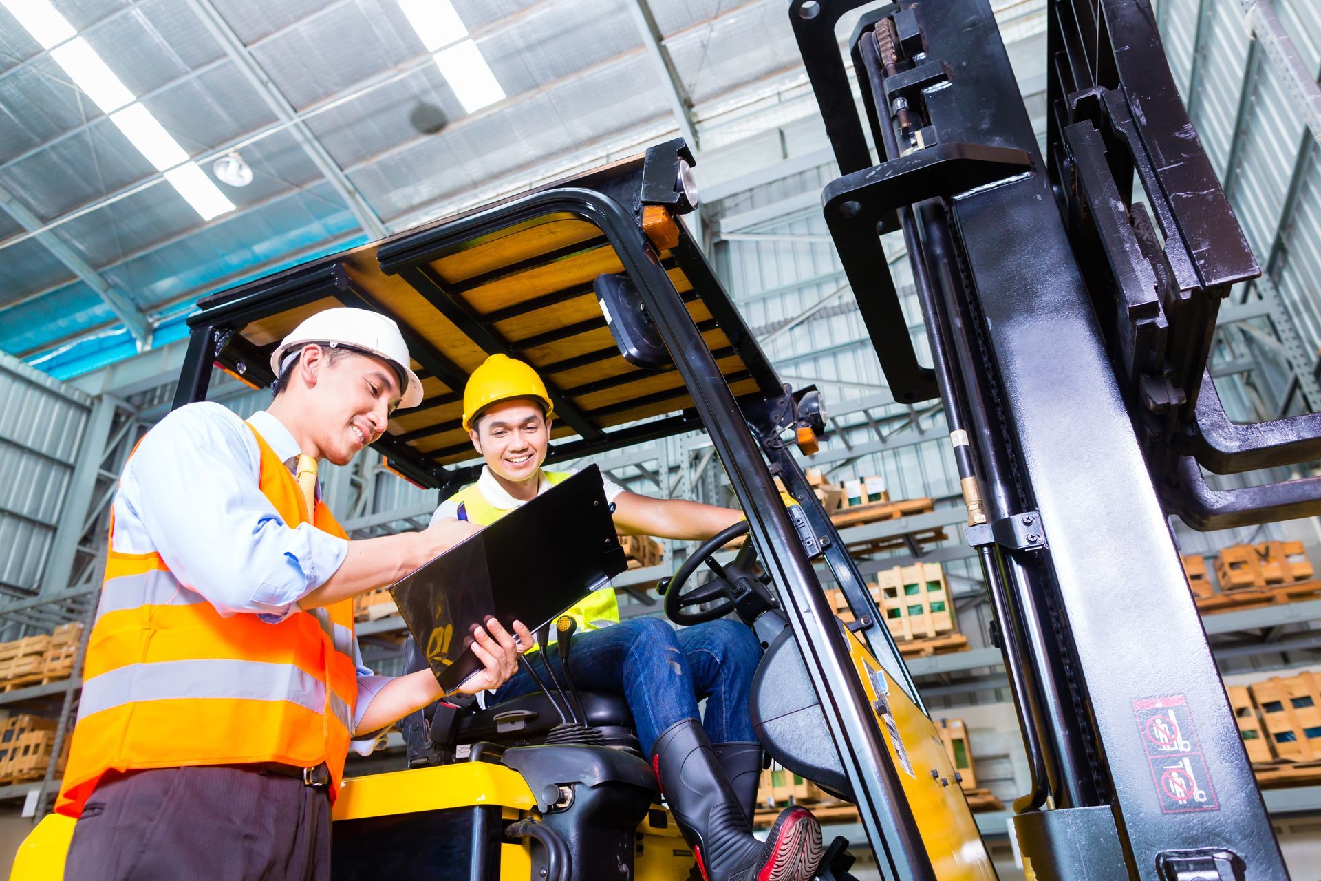 Two men in hard hats and vests reviewing paperwork near a forklift in a warehouse.