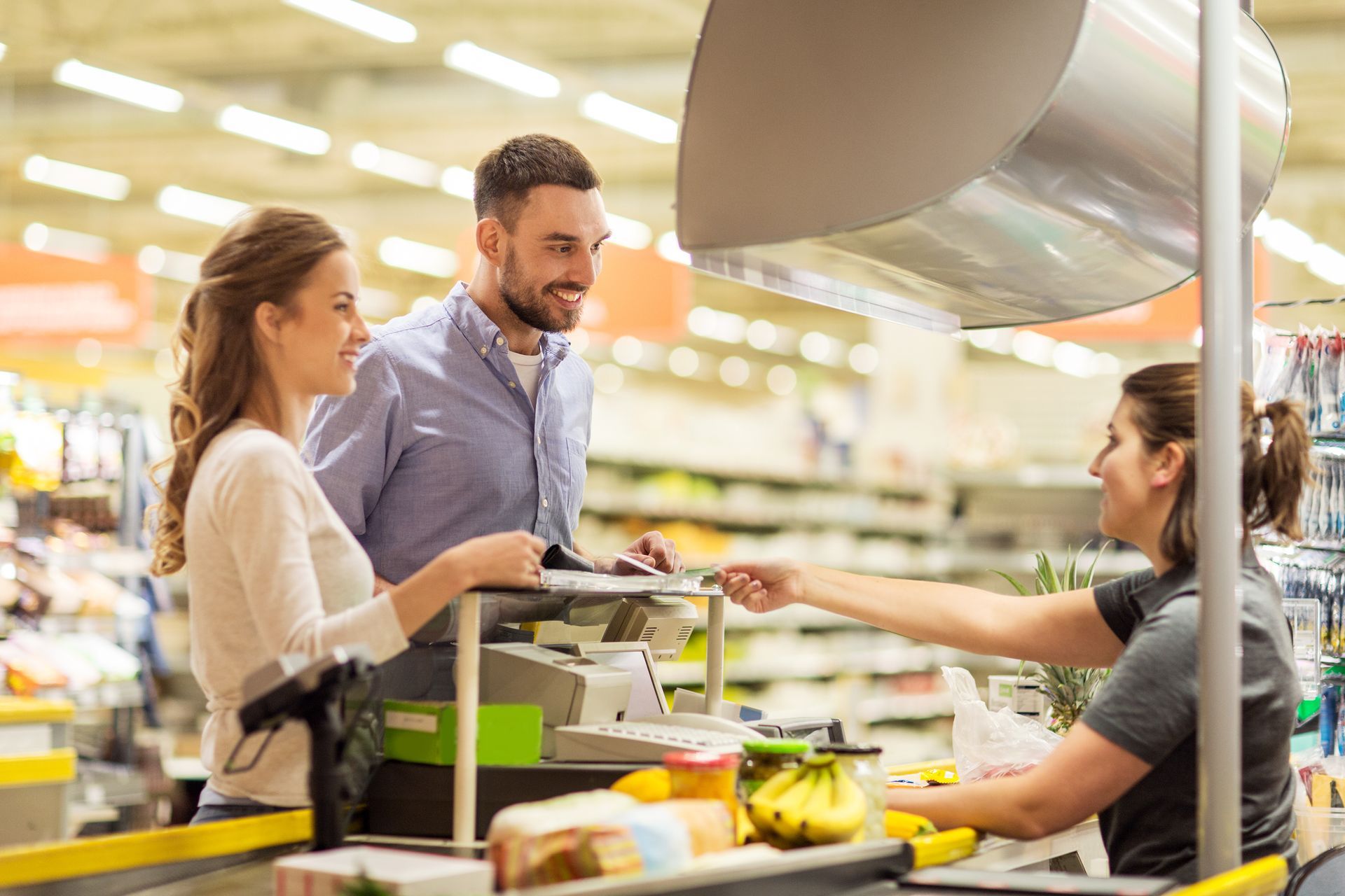 Couple paying for groceries at a checkout counter; smiling cashier hands them a card.