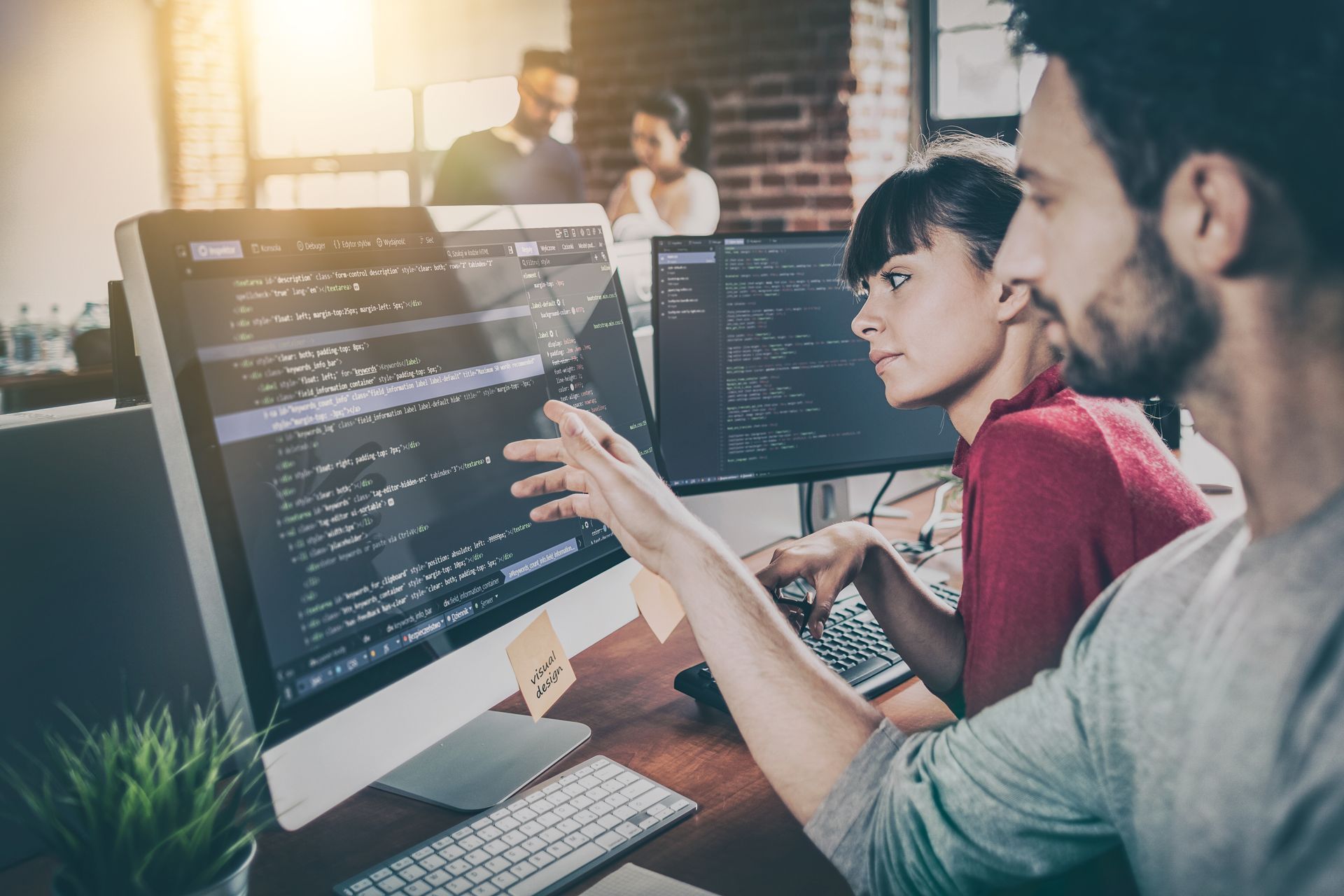 Two people coding on computer screens in a modern office, one pointing at the code.