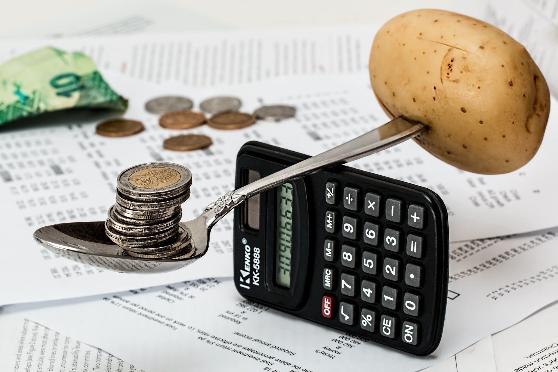 A calculator, coins, and a potato are balanced on a spoon, with money and receipts in the background.