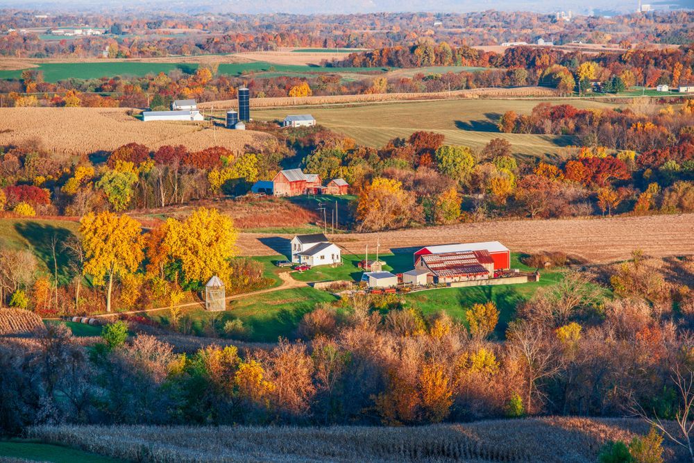 Rolling farmland with fall foliage, barns, and silos under a blue sky.
