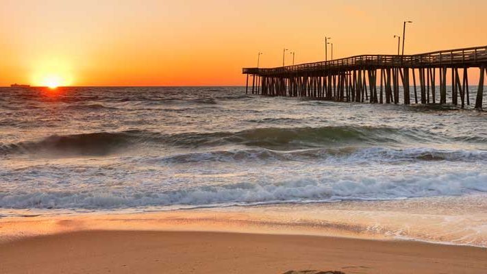 Sunset over ocean, pier in silhouette. Warm orange sky, waves crashing on sandy beach.