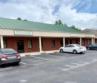 Brick strip mall storefront with green roof, parked cars, and cloudy sky