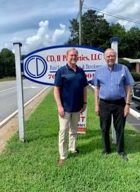 Two men standing beside a roadside business sign on a grassy shoulder under a cloudy sky