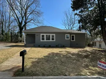 Single-story gray house with a front lawn, mailbox, and for-sale sign under a clear blue sky.