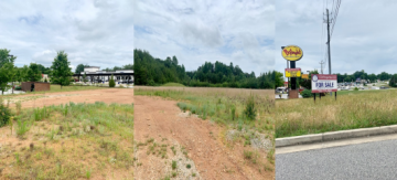 Rural roadside with grassy field, dirt path, and a distant yellow sign near a highway.