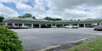 Strip mall storefronts and parked cars along a mostly empty parking lot under cloudy skies