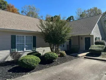 Single-story beige house with shrubs and a paved driveway under a clear blue sky