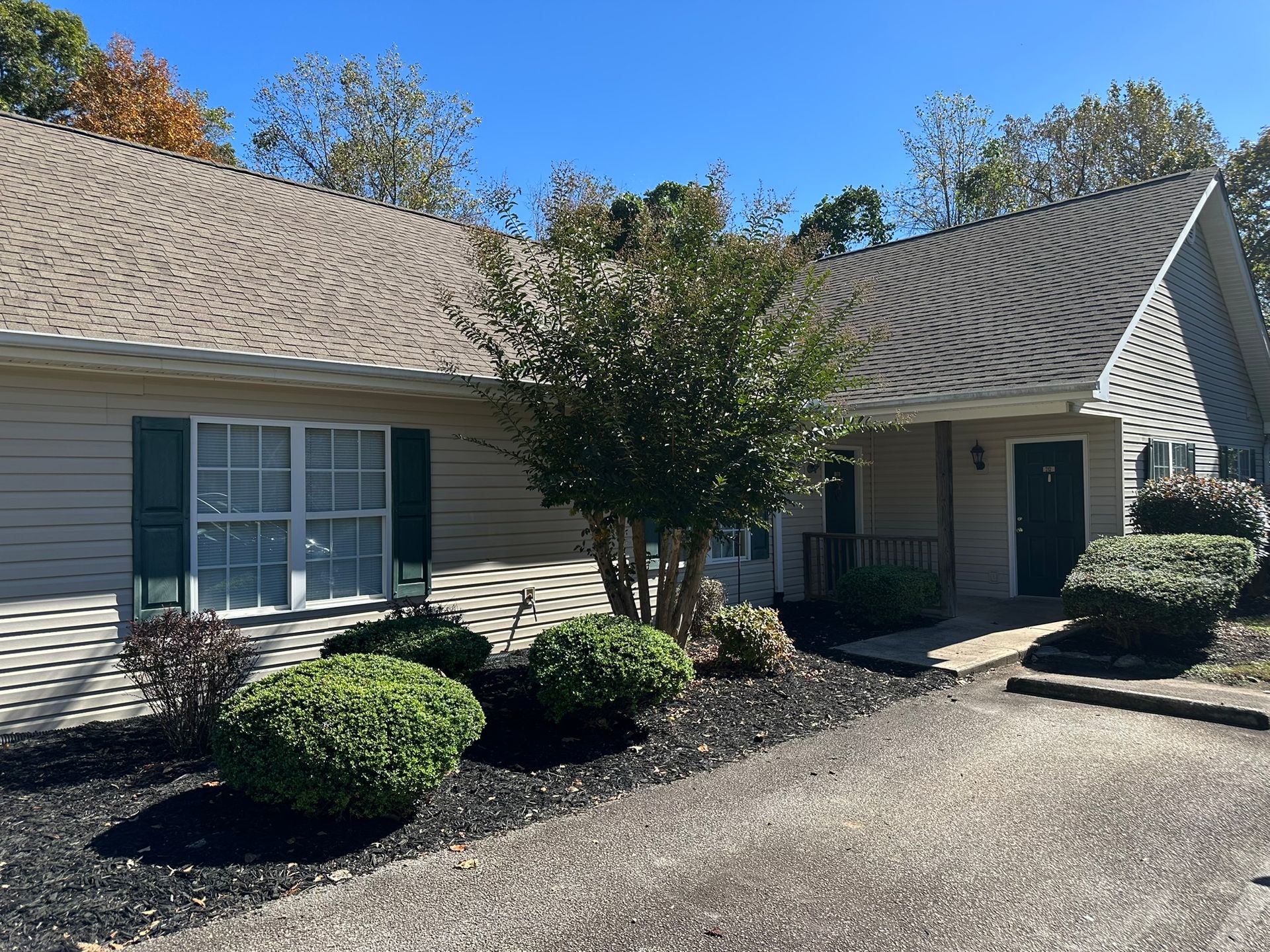 Single-story house exterior with gray siding, green shutters, shrubs, and a gravel driveway under a blue sky
