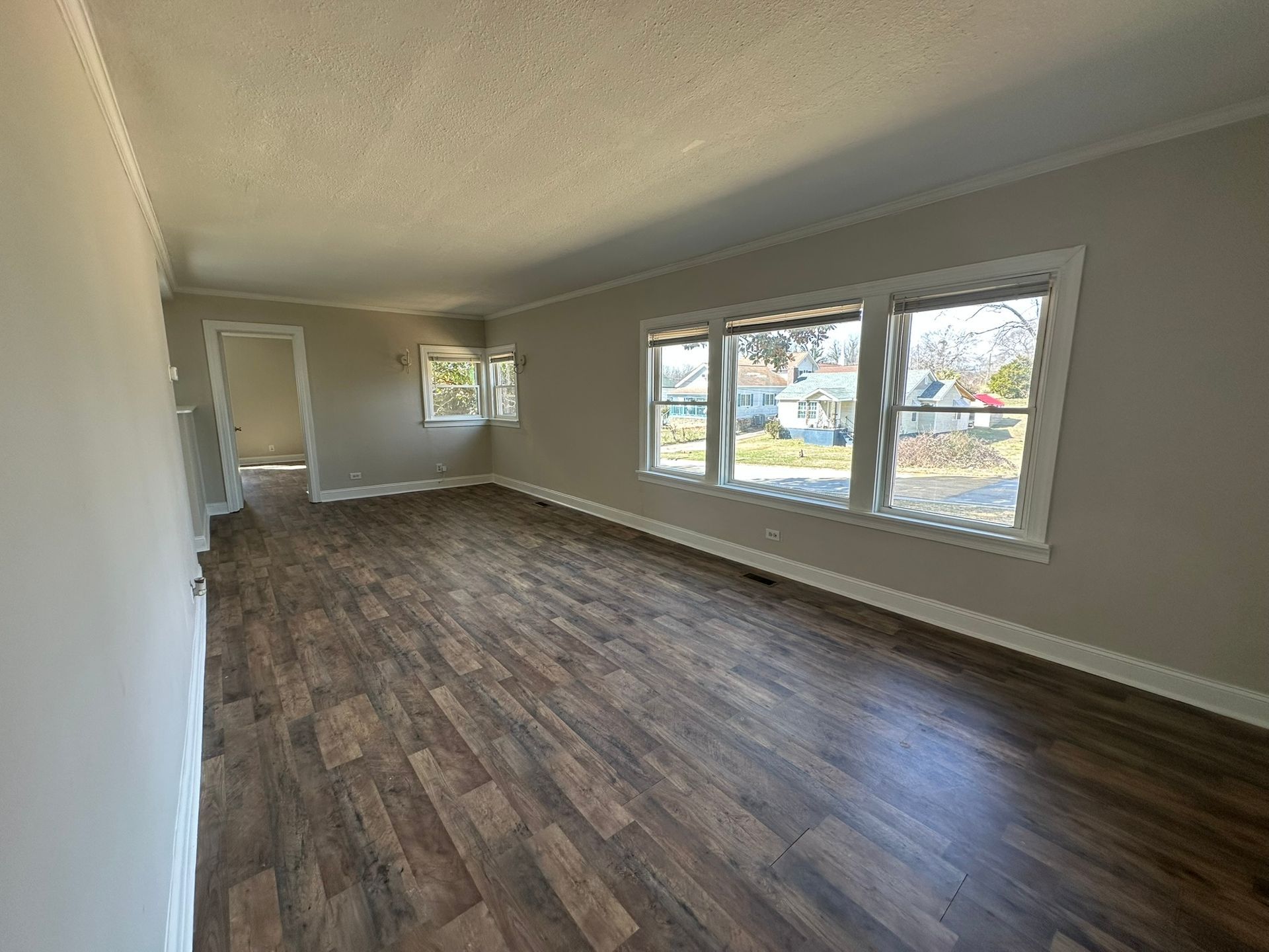 Empty living room with wood-look floors, white walls, and large windows along one side.