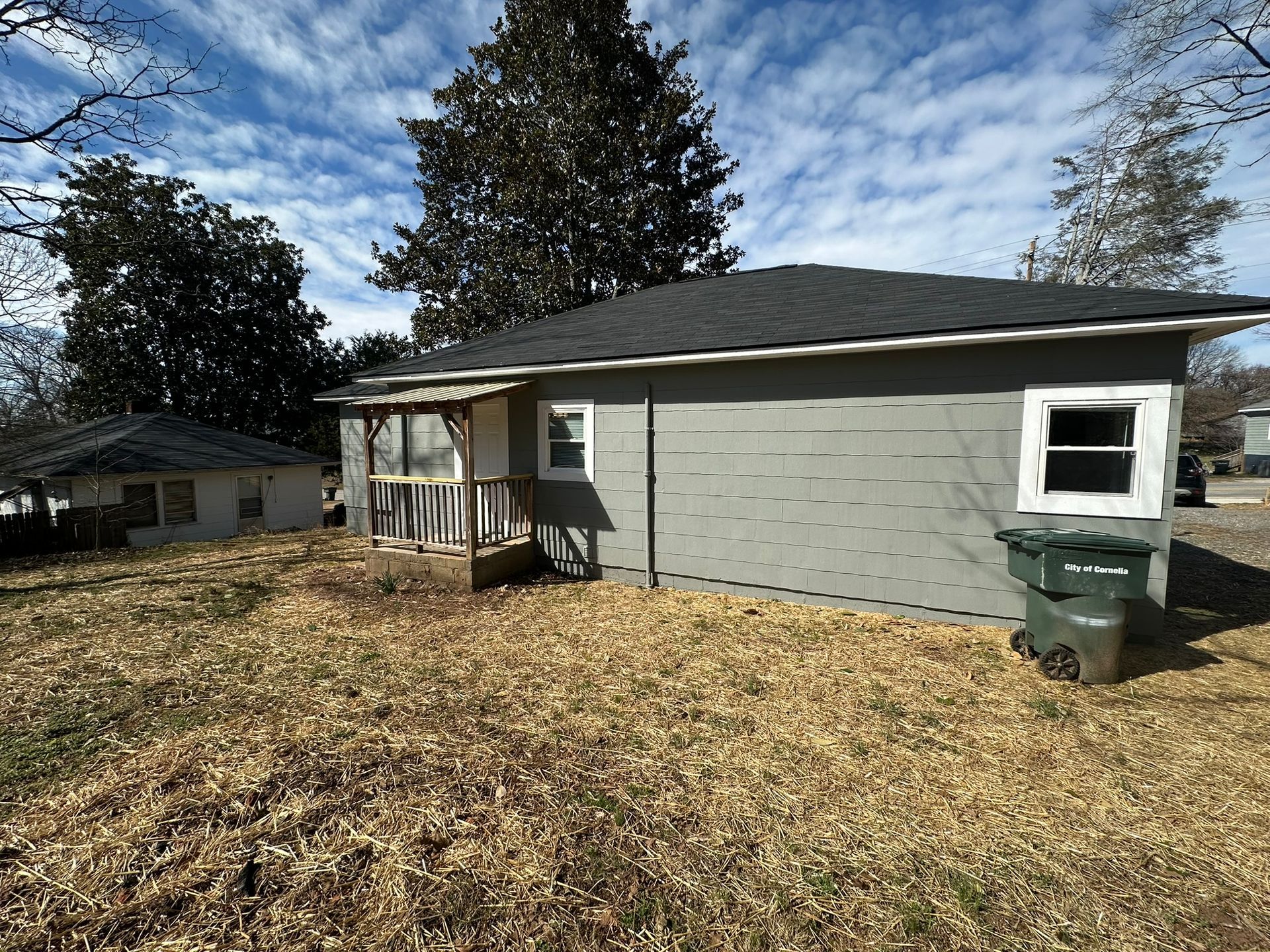 Gray house with black roof, small porch, and two trash bins in a leaf-covered yard under a cloudy sky