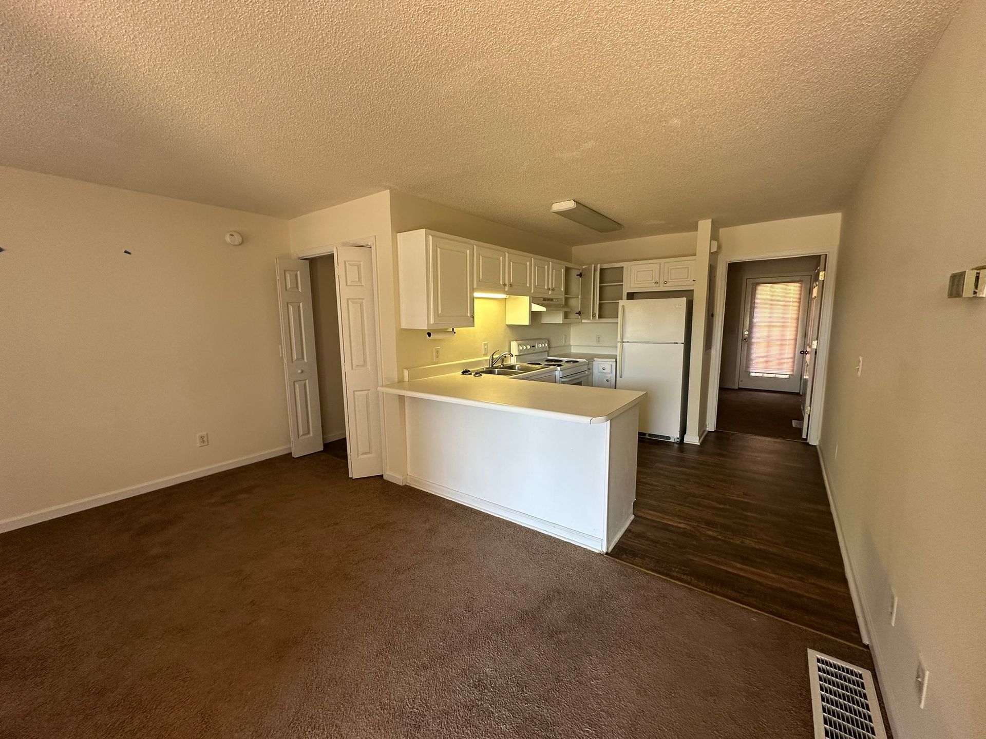 Empty living room with brown carpet, white kitchen cabinets, and a breakfast bar.