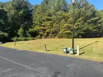 Parking lot edge with a grassy embankment, trees, a lamppost, and two utility boxes on a sunny day