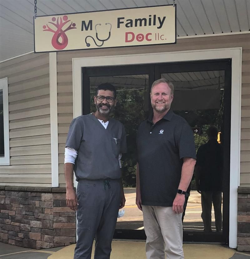 Two men stand outside a family doctor office sign, one in scrubs and one in a black polo.