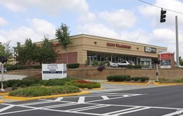 Street view of a shopping center with storefronts, landscaped median, and traffic lights at an intersection.